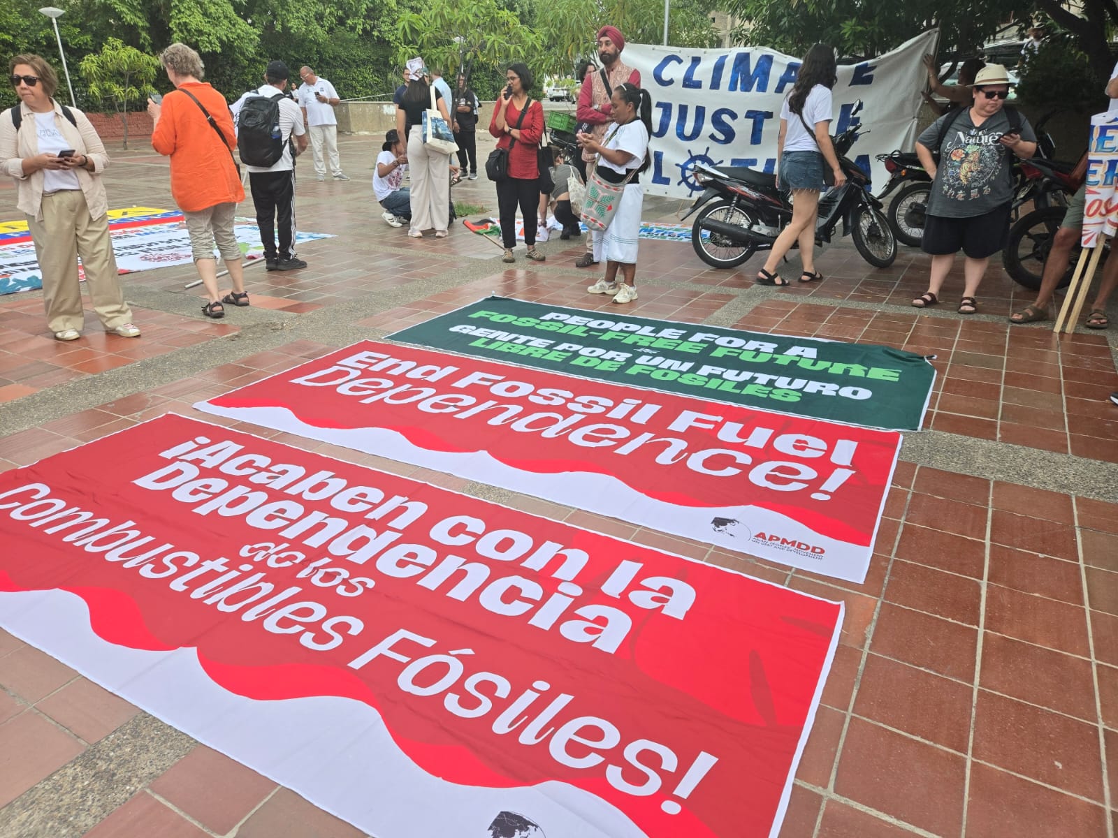 Fotografia mostra cartazes que discutem transição energética limpa em três línguas: espanhol, inglês e português. A frase nos cartazes é: chega da dependência de combustíveis fósseis. Pessoas estão ao fundo dos cartazes. 